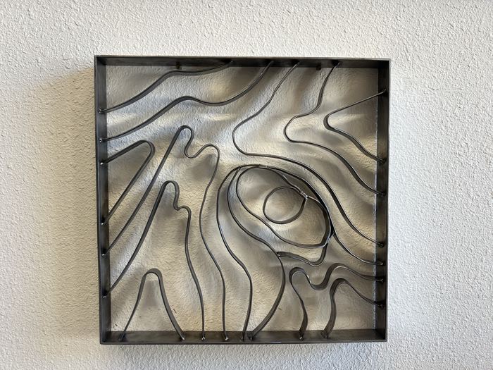 Square Topograin wall art in raw steel with a wood-knot form, hung on a white textured wall
